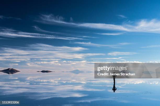 lone, silhouetted traveler in safari hat stops to admire the beauty of the salt flats which are reflecting the clouds and mountains after rainfall, uyuni, bolivia - cloud sales fotografías e imágenes de stock