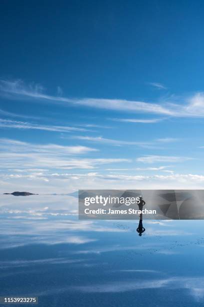 lone, silhouetted traveler in safari hat stops to admire the beauty of the salt flats which are reflecting the clouds and mountains after rainfall, uyuni, bolivia - cloud sales fotografías e imágenes de stock