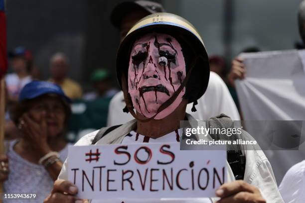 July 2019, Venezuela, Caracas: "SOS - Intervention", is written on the poster of a masked demonstrator at a rally against the government of the head...
