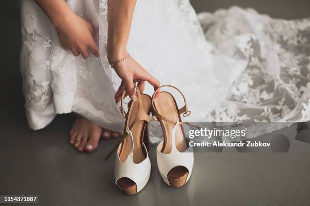 the bride, girl or young woman in a beautiful elegant modern stylish wedding dress reaches for light fashionable high-heeled shoes to put on, close-up. the day of the wedding or the morning. - girls high heels stock-fotos und bilder