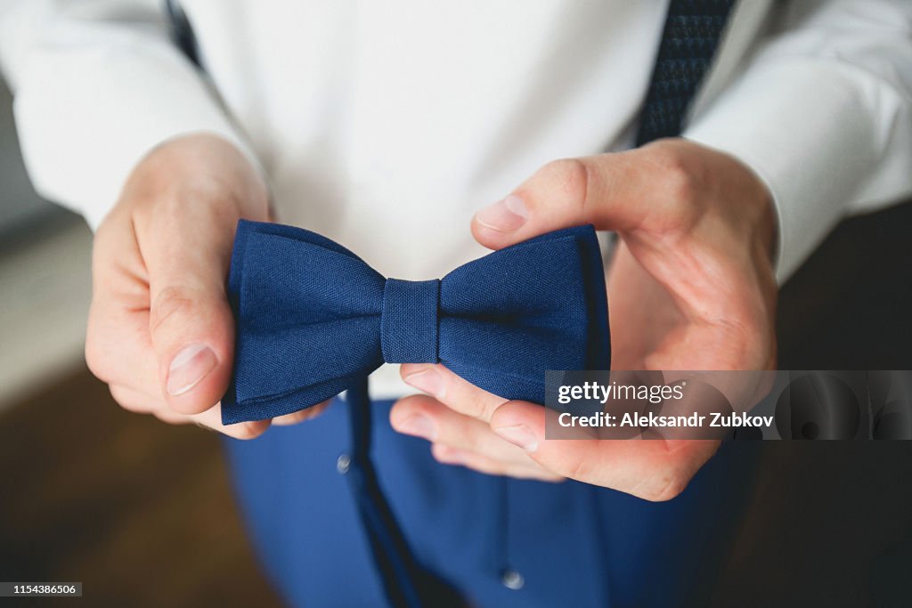 A man, a groom or a businessman in a stylish suit holding a blue bow tie, a fashion accessory. Preparing the groom for the wedding day. Blurred background, close-up.