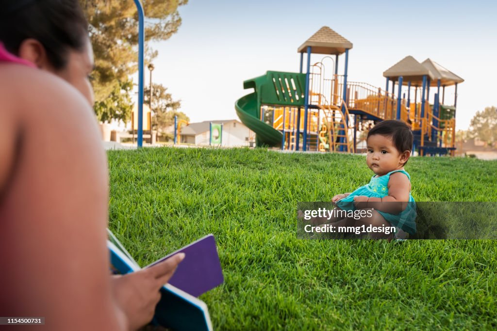 A woman holding a childrens book tries to engage the attention of a baby girl sitting in the lawn of a kids playground.