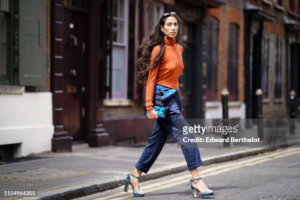 Ciinderella Balthazar wears an orange turtleneck pullover, striped cropped pants, a blue geometric pattern clutch, blue shoes, sunglasses, during...