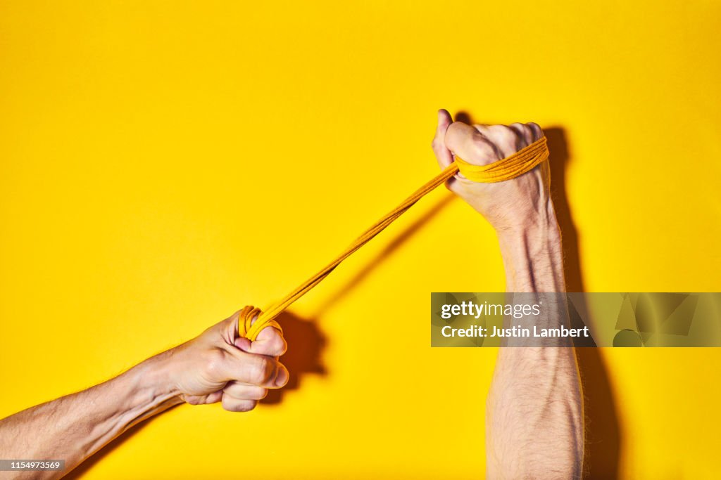 Hands with cord wrapped around pulling apart symbolising tension and pulling, on a bright yellow backdrop