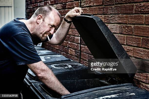 mature male vagrant smokes, rummaging in refuse bin - wheelie bin stock pictures, royalty-free photos & images