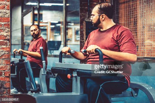 ein übergewichtiger mann in einem fitnessstudio mit dem übungsgerät beim blick auf den spiegel - trainingsmaschine stock-fotos und bilder