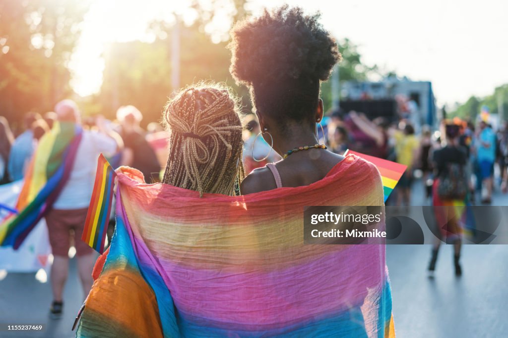 Pares fêmeas novos que abraçam com o lenço do arco-íris no evento do orgulho