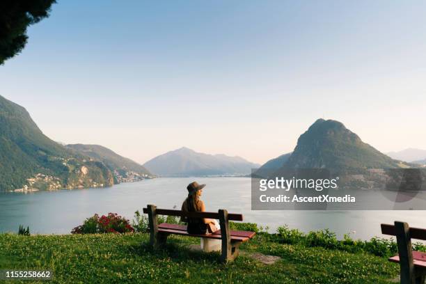 woman relaxes above lake and mountains on bench - lugano switzerland stock pictures, royalty-free photos & images