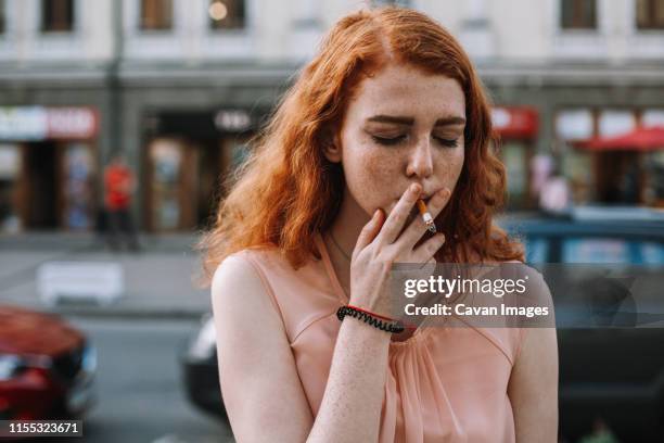 young woman with freckles smoking cigarette while standing in street - young woman red hair urban stock pictures, royalty-free photos & images
