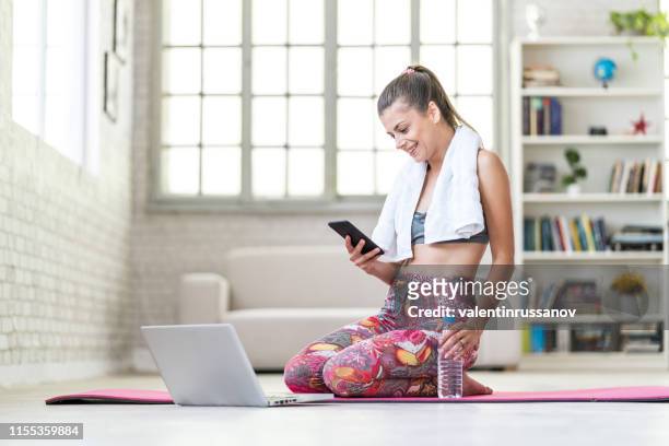 woman using smart phone on exercise mat in front of her laptop - exercise computer stock pictures, royalty-free photos & images