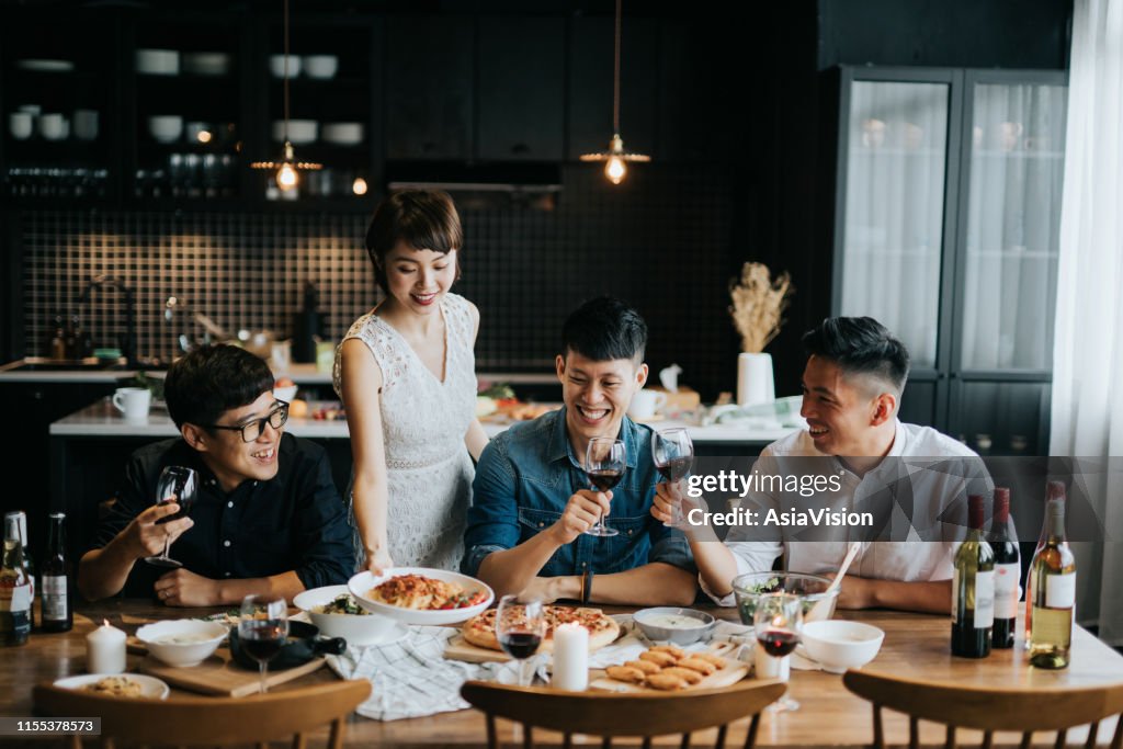 Smiling young Asian woman serving food to friends during party at home, they are smiling joyfully and toasting with red wine