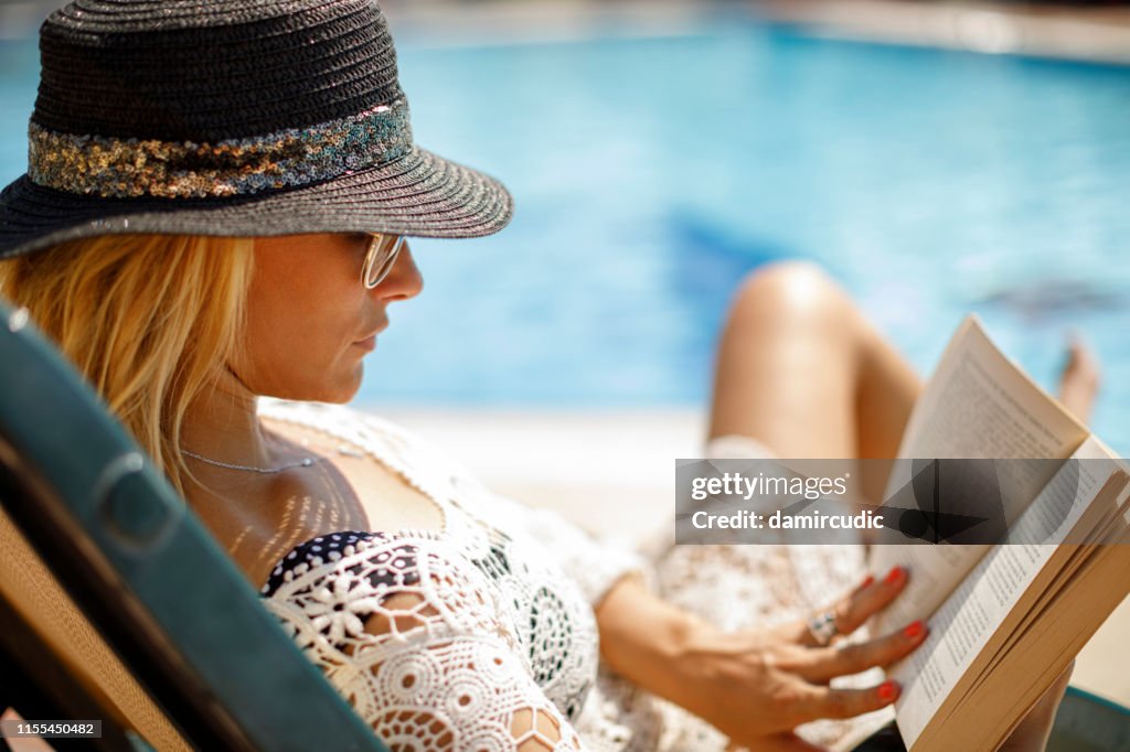 Frau mit Hut und Sonnenbrille beim Lesen eines Buches in ihrem Hinterhof