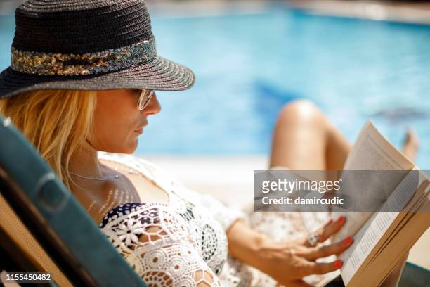 donna con cappello e occhiali da sole con la lettura di un libro nel suo cortile - sdraio da spiaggia foto e immagini stock