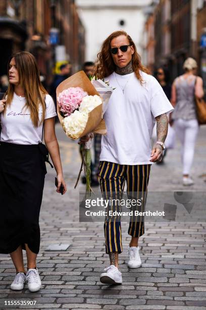 Guest wears sunglasses, a white T-shirt, black cuffed pants with yellow stripes, white sneakers, and holds a bundle of hydrangeas flowers, during...