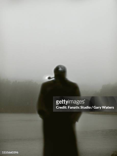 an atmospheric image of a man from the back in a heavy overcoat smoking a pipe in the rain in front of a lake - grey overcoat stock illustrations