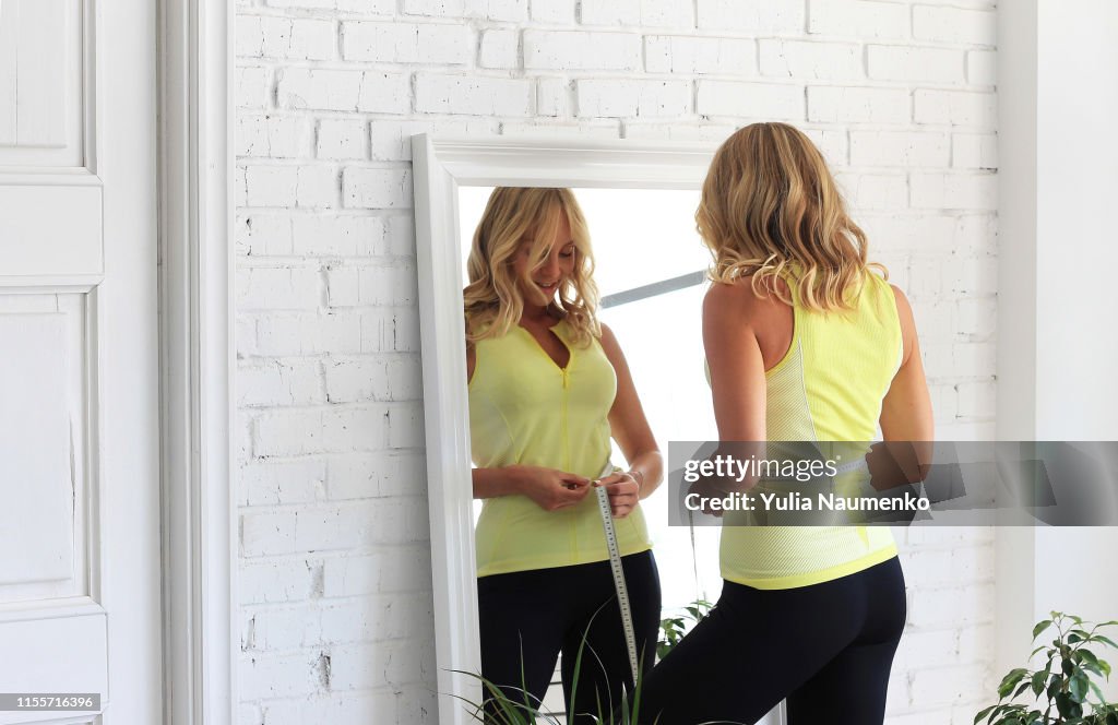 Stay in shape. Young woman with athletic body measures her waist with a measure type in front of a mirror.