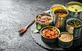 Various open tin cans of canned food on a stone Board with a fork.