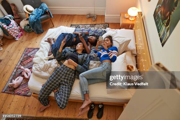 female friends using phones while relaxing on bed - three people in bed stock pictures, royalty-free photos & images