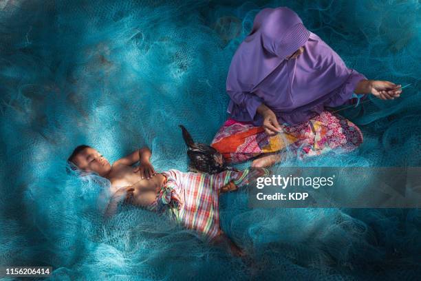 portrait of muslim woman is repairing fishing nets while her son beside sleeping - south east asia stock-fotos und bilder