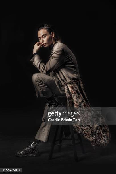 un hombre chino asiático sentado en un taburete con ropa de moda mirando fresco a la cámara con negro fondo estudio disparar - male fashion fotografías e imágenes de stock