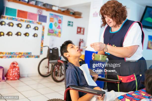 latina nurse in day care feeding boy with celebral palsy in wheelchair. - learning disability nurse stock pictures, royalty-free photos & images