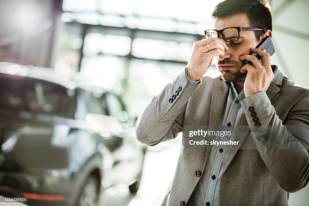Geschäftsmann kopfschmerzen, während er auf dem Handy in einem Auto-Showroom spricht.