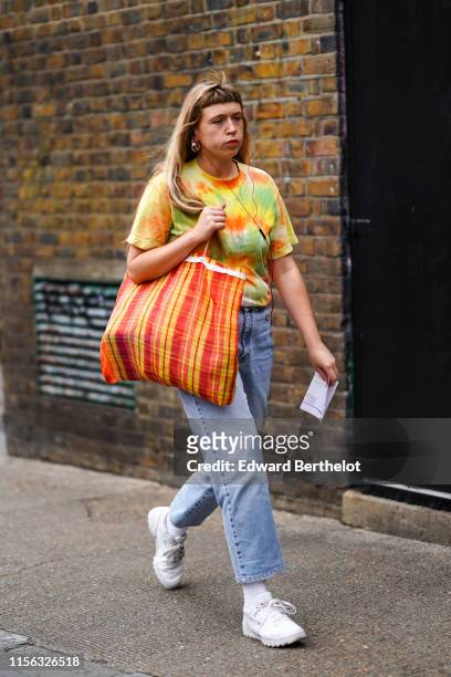 Guest wears earrings, a green, yellow and orange tie and dye t-shirt, jeans, a red and yellow checkered large tote bag, white Reebok sneakers, during...