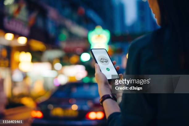 Over the shoulder view of woman using mobile app on smartphone to arrange taxi ride in downtown city street, with busy traffic scene as background