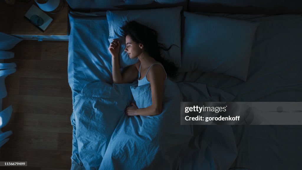 Top utsikt över vacker ung kvinna sova Cozily på en säng i hans sovrum på natten. Blå nattliga färger med kallt svagt Lamppost ljus lyser genom fönstret.
