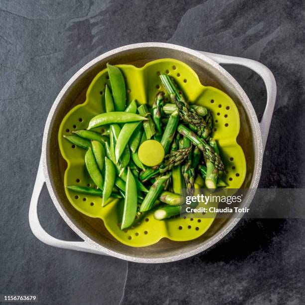 steamed asparagus and sugar snap peas on a sieve in a cooking pot on black background - steamed stock-fotos und bilder