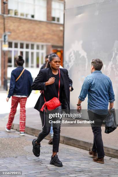 Guest wears sunglasses, a black top, a black jacket, black leather pants, black Balenciaga sneakers, a red Gucci bag, during London Fashion Week...