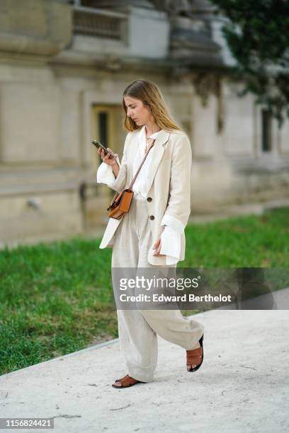 Guest wears a white shirt, a cream-color pantsuit with wide-legs pants, a tan-color leather bag, tan -color flat mules, outside AMI, during Paris...