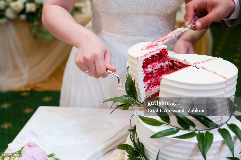 The bride and groom cut an elegant three-tiered white wedding cake decorated with natural flowers or roses and green leaves on a white wooden table.