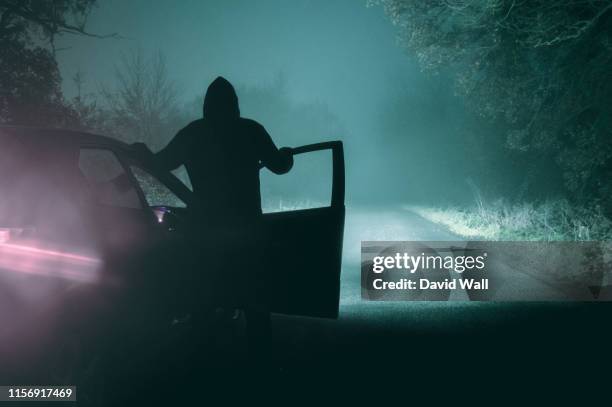 a lone, hooded figure standing next to a car looking at an empty misty winter country road silhouetted at night by car headlights - dark forest stock-fotos und bilder