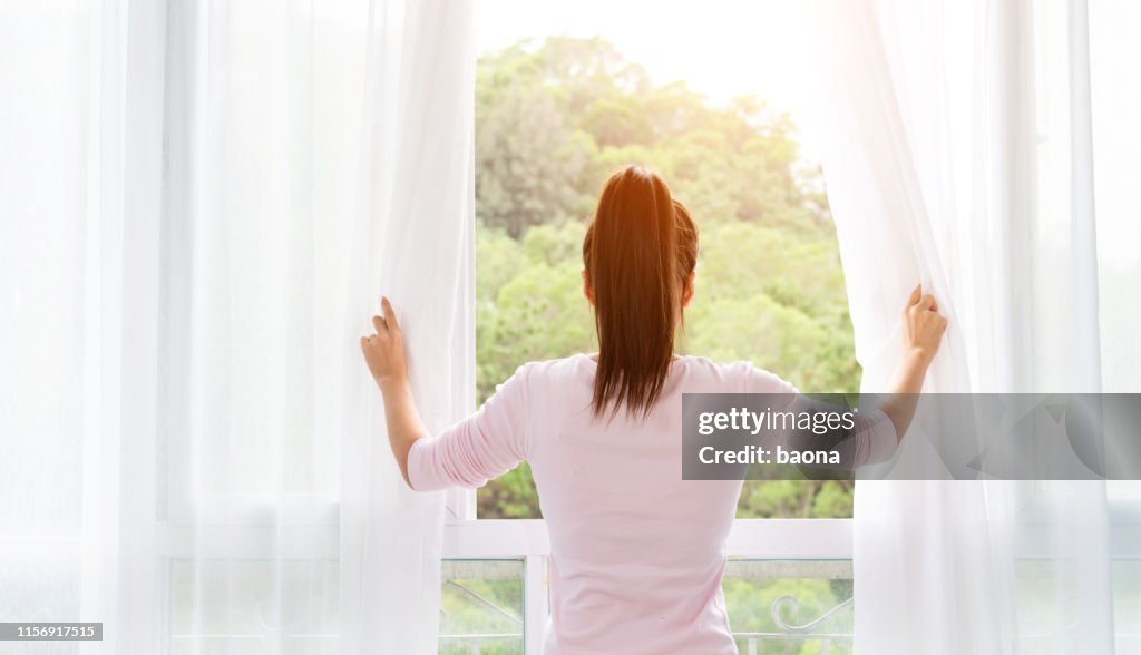 Asian young woman opening the curtains and looking out of window