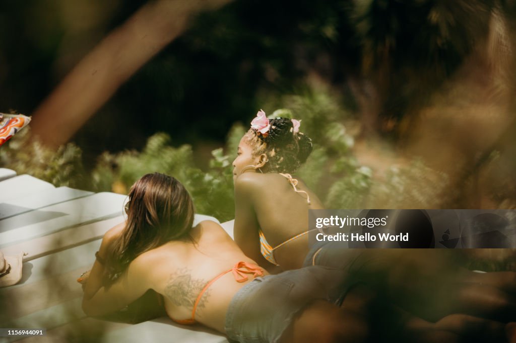 Girlfriends sunbathing at poolside seen through foliage