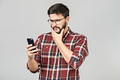 Puzzled bearded man with confusion reading shocking news or browsing newsfeed via social networks using mobile phone