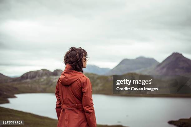 young woman stands on mountain looking at view of loch in scotland - wandern regen stock-fotos und bilder