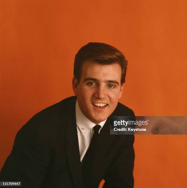 Headshot of Billy J. Kramer, British singer, wearing a black jacket, white shirt and black tie, against an orange background, circa 1963.