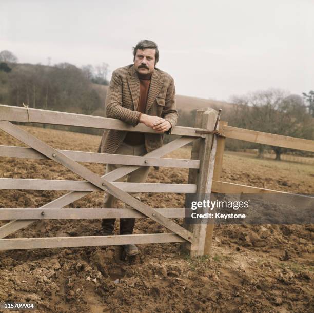 Oliver Reed , British actor, wearing a tweed jacket and wellington boots while leaning on a five-bar gate in a field, Great Britain, circa 1975.