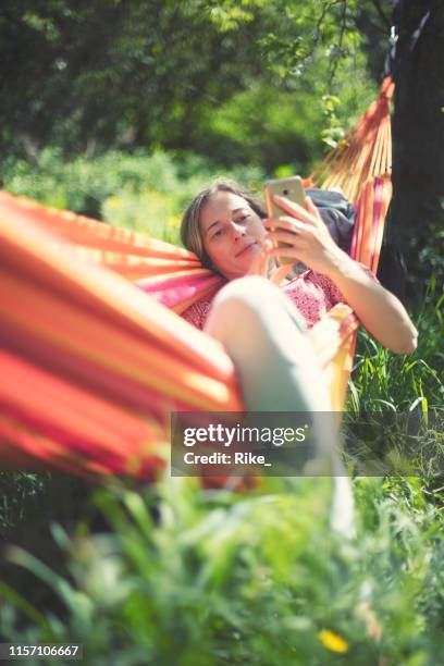 jonge vrouw ontspant in hangmat op een mooie zomerdag en kijkt in haar smartphone - hammock stockfoto's en -beelden