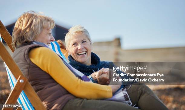 smiling lesbian couple sitting and talking on beach - man on the beach relaxing in deckchair stock-fotos und bilder