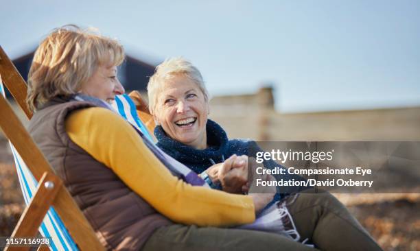 smiling lesbian couple sitting and talking on beach - gay seniors fotografías e imágenes de stock