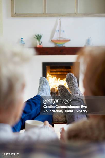 mature lesbian couple with hot drinks on living room sofa at fireside - hot wife stockfoto's en -beelden