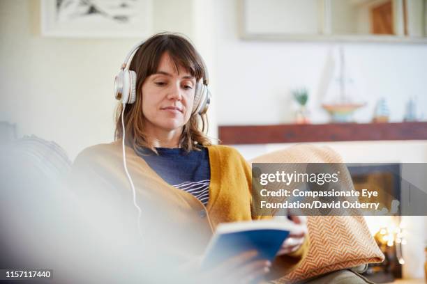 woman with headphones reading in living room - generation x stock-fotos und bilder