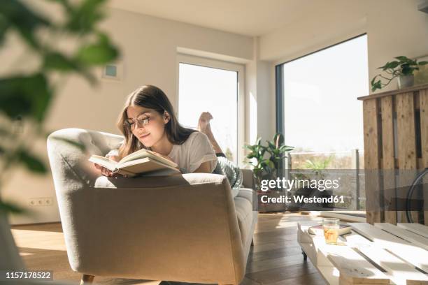 relaxed young woman lying on couch reading a book - escapism reading stock pictures, royalty-free photos & images