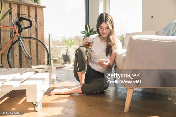 young woman sitting on the floor at home with drink and cell phone - bel appartement stockfoto's en -beelden