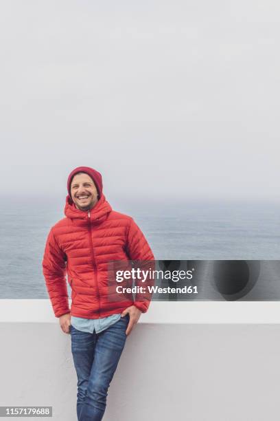 portugal, algarve, sagres, cabo de sao vicente, happy man standing at a wall above the sea - puffer jacket photos et images de collection