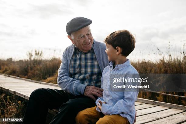 happy grandfather sitting with his grandson on boardwalk looking at each other - season 6 stock-fotos und bilder