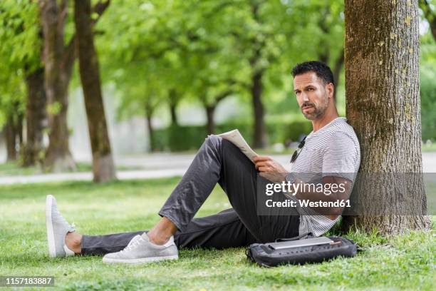 man leaning against a tree in park reading newspaper - leaning tree stock pictures, royalty-free photos & images